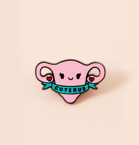Pink enamel smiling cartoon uterus pin features hearts at the end of its fallopian tubes and is wrapped in a blue banner that says, "Cuterus"