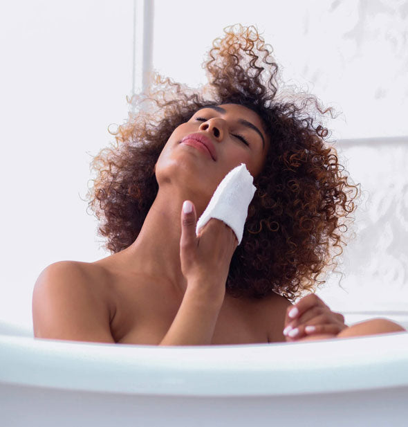 Model sitting in a bathtub demonstrates use of the Daily Facial Micro Scrubber on face