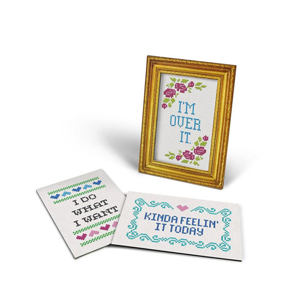 Cross-stitch artwork in gold frame says, "I'm over it." flanked by purple flowers and leaves; two other cross-stitch design lay nearby which read, "I do what I want" and "Kinda feelin' it today"