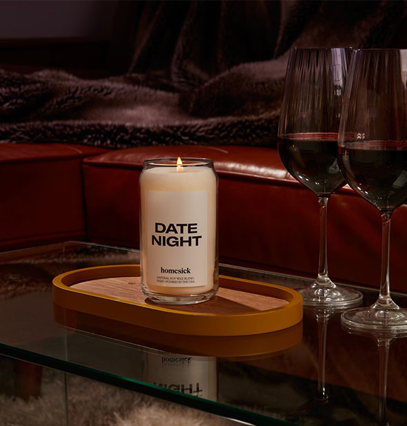 A lit Date Night candle sits on a wood and glass surface in a darkened room next to two wine glasses