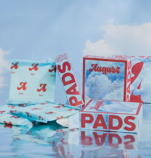 Grouping of August brand day pads in wrappers and box packaging on a backdrop of blue sky with clouds