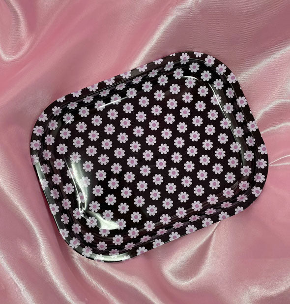 Rectangular black tray with rounded corners and all-over daisy pattern sits on a pink satin cloth