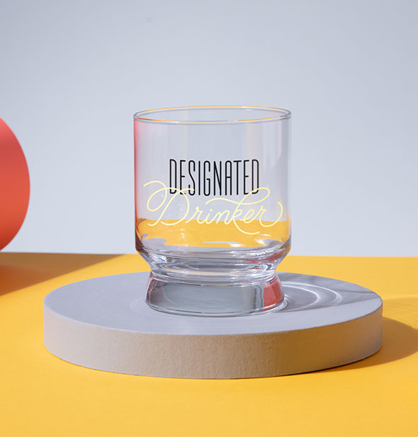 Designated Drinker rocks glass sits atop a round blue pedestal on colorful backdrop