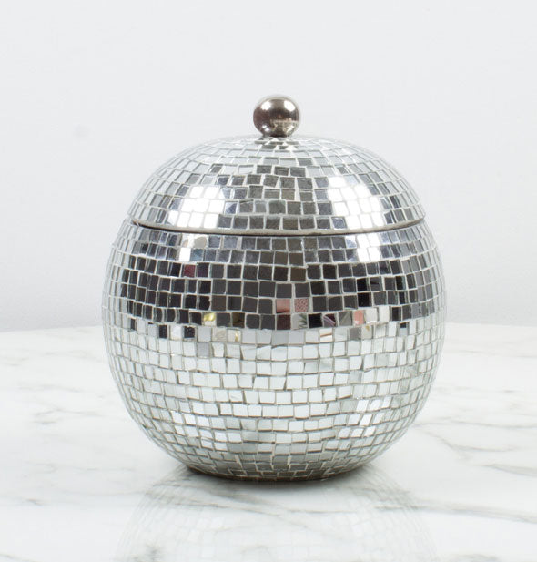 Round container sitting on a marble surface has an all-over mirrored tile finish and lid topped with a spherical handle