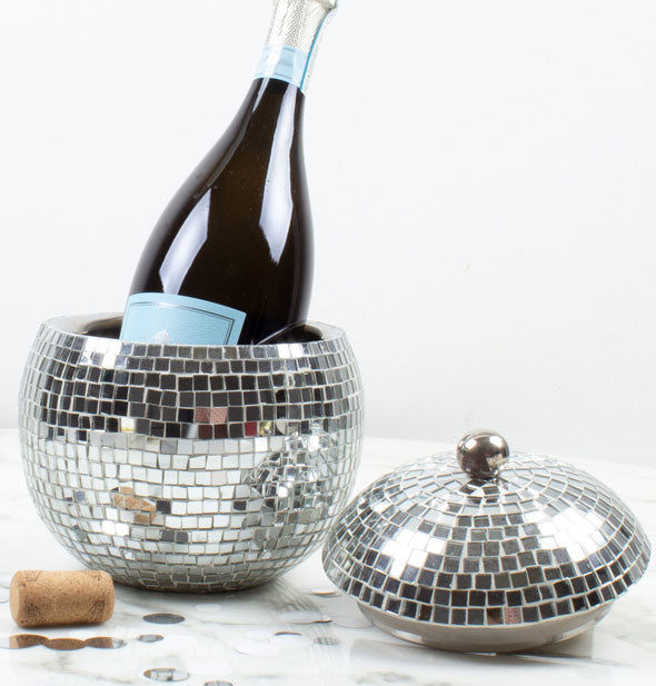 Champagne bottle sits in an opened disco ball ice bucket; the lid, a cork, and silver confetti lay to the side