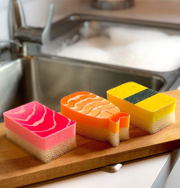 Tuna, shrimp, and tomago dish sponges on a wooden cutting board propped on a sink's edge