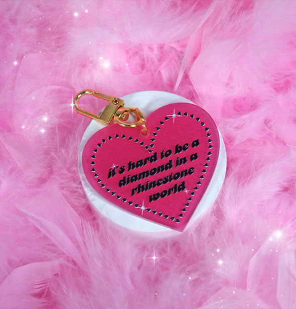 Pink heart-shaped keychain with gold clasp resting on a round white pedestal on pink feathers says, "It's hard to be a diamond in a rhinestone world" in black italic lettering