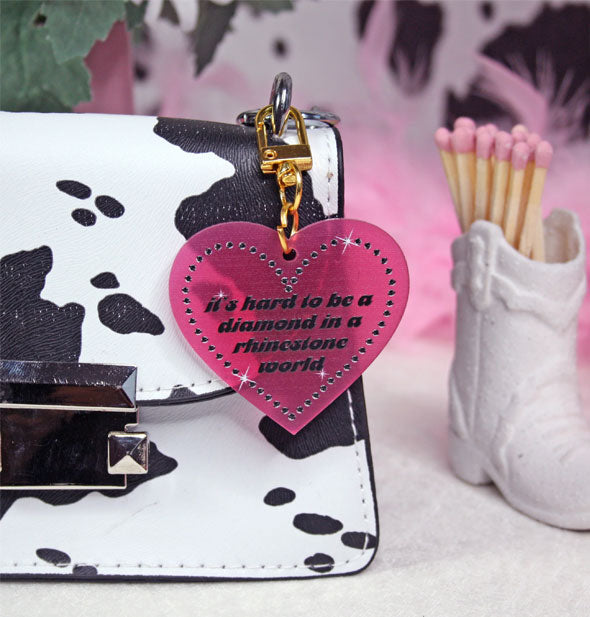 Translucent pink heart-shaped keychain with gold hardware attached to a black and white cowhide print handbag next to a small white cowboy boot match holder says, "It's hard to be a diamond in a rhinestone world"
