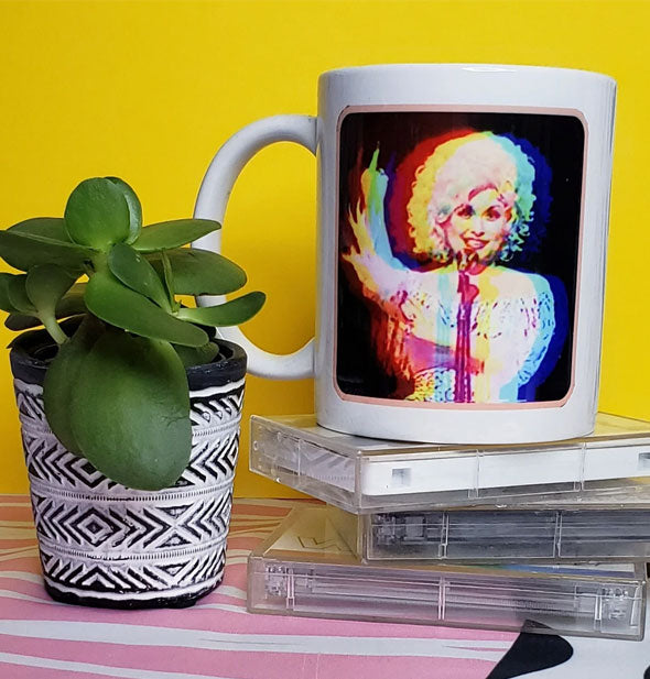 Coffee mug with distorted image of Dolly Parton appearing to raise her middle finger sits on a stack of cassette tapes next to a potted succulent