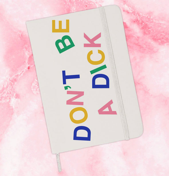 White journal with elastic band and ribbon place marker on a pink marbled background says, "Don't be a dick" in multicolor lettering