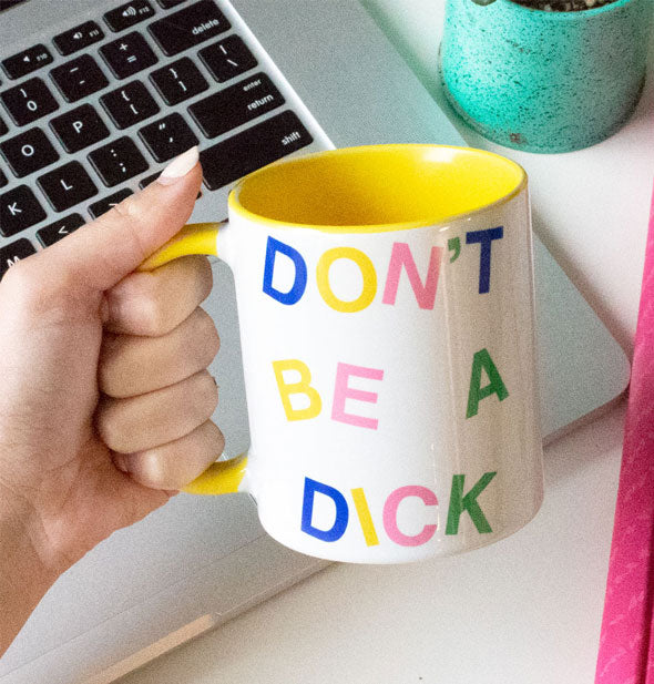 Model's hand holds a Don't Be a Dick mug in front of a laptop keypad