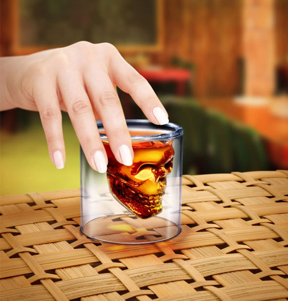 Hand holds a skull shot glass filled with brown liquor.