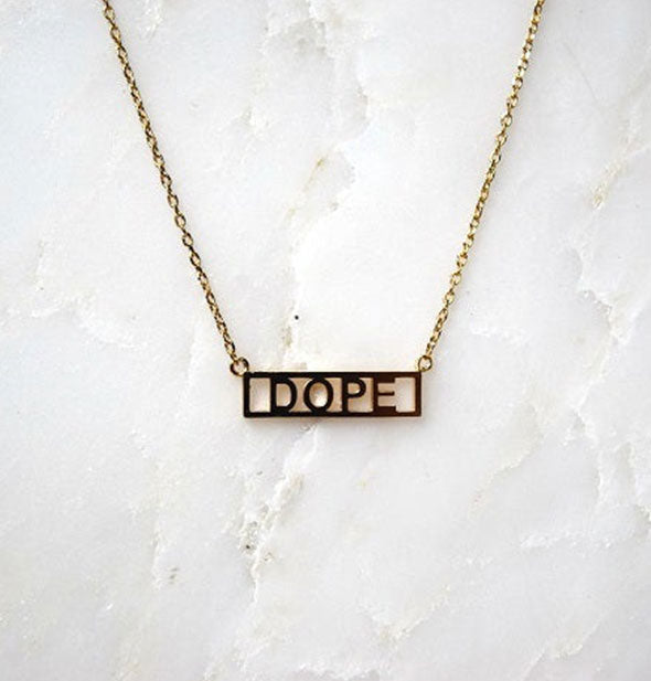 Gold bar necklace on white marble surface says, "Dope"