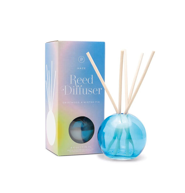 Round blue glass reed diffuser with five wooden reeds sticking out of it sits next to box packaging