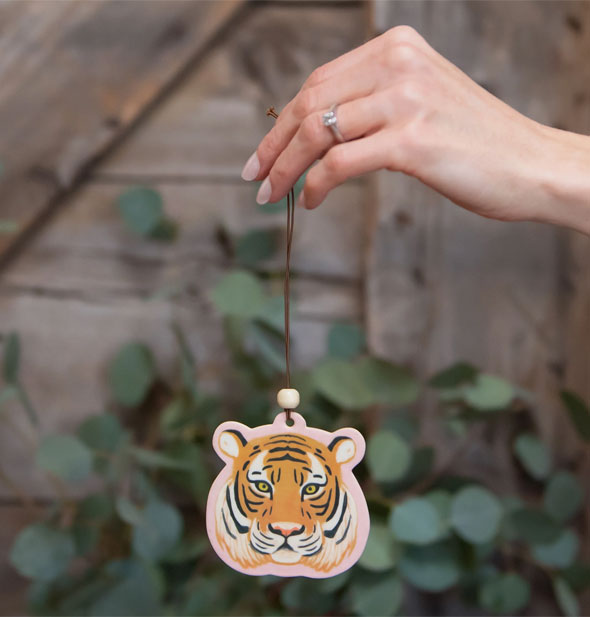 Model's hand holds a tiger head car air freshener by its string against a botanical and wood backdrop