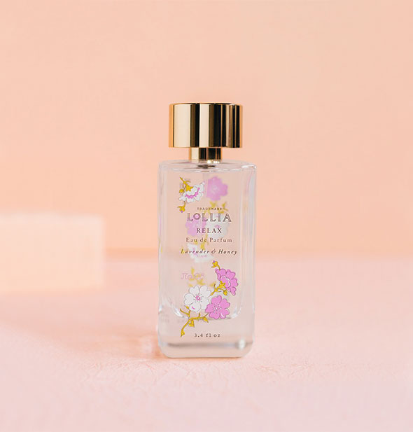 Rectangular clear glass bottle of Lollia Relax Eau de Parfum with pink and white floral design and gold cap is set against a pink backdrop