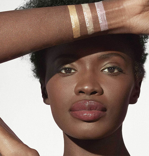 Model with dark skin tone raises arm painted with three labeled shades of Jane Iredale Glow Time Highlighter Stick