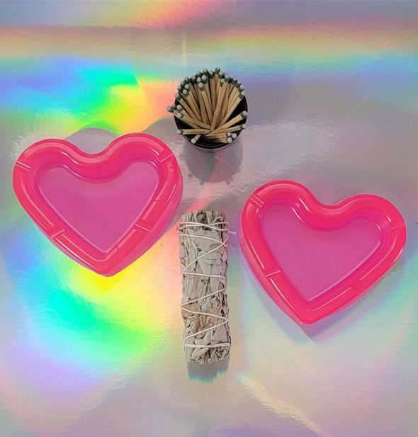 Two pink heart-shaped ashtrays staged with matches and a sage bundle on holographic surface