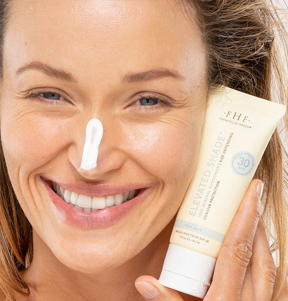 Smiling model with a stripe of product applied to nose holds a bottle of FarmHouse Fresh Elevated Shade 100% Mineral Sunscreen