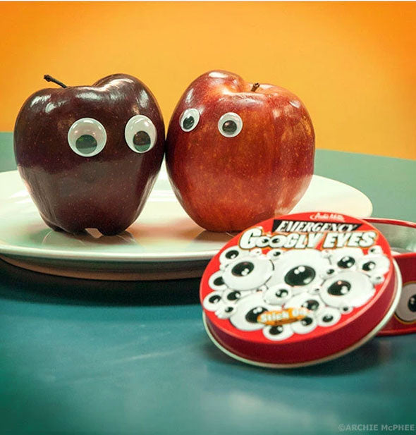 A lid to an Emergency Googly Eyes tin lays in front of two apples with googly eyes applied to them