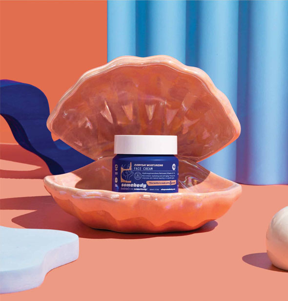 Blue and white pot of Somebody Everyday Moisturizing Face Cream sits in a ceramic coral-colored oyster shell on a matching backdrop staged with blue structural shapes