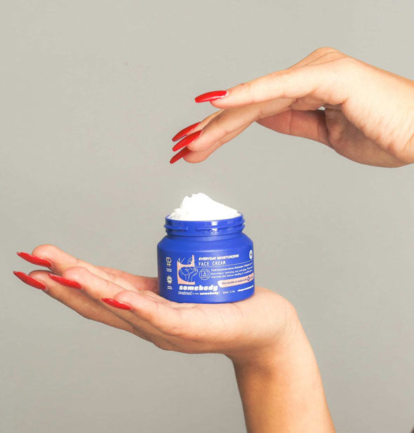 A blue pot of Somebody Everyday Moisturizing Face Cream rests in the palm of a model's hand with the other hand reaching over top
