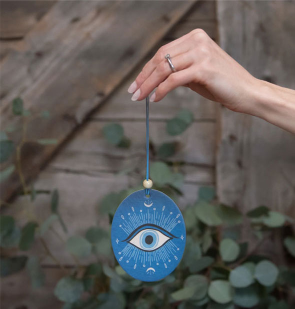 Model's hand holds a rounded blue car air freshener featuring an evil eye design against a wood and botanical backdrop