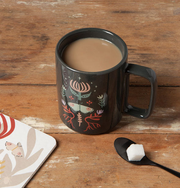Moth mug filled with coffee sits on a wooden tabletop near a spoon and sugar cube
