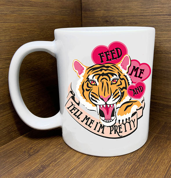 White coffee mug with roaring tiger illustration flanked by pink hearts and banner printed with the words, "Feed me and tell me I'm pretty" sits on a wooden backdrop