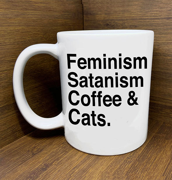 White coffee mug says, "Feminism Satanism Coffee & Cats" in black lettering and sits on a wooden backdrop