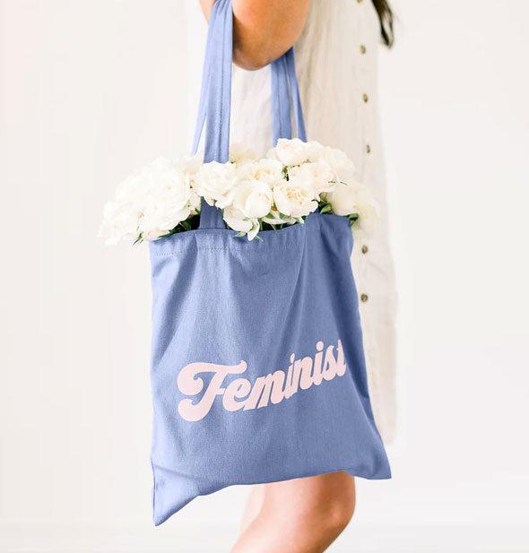 Model holds a blue Feminist tote bag filled with white flowers in the crook of her arm