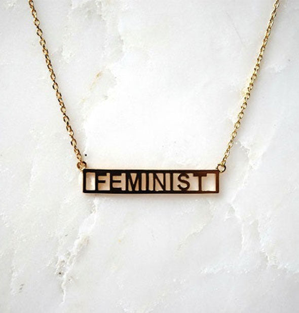 Gold bar necklace on white marble surface says, "Feminist"