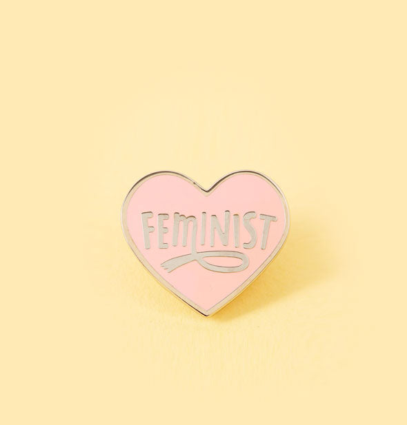 Pink enamel heart-shaped pin says, "Feminist" with loop accent on the M