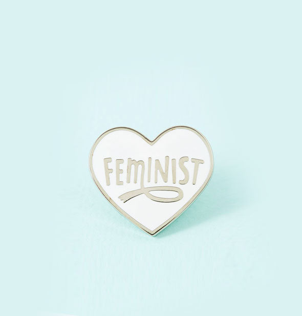 White enamel heart-shaped pin says "Feminist" with loop accent on the M