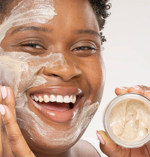 Smiling model applies cleansing polish to face with one hand and holds a jar of product in the other