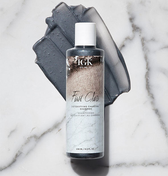 Bottle of IGK First Class Detoxifying Charcoal Shampoo with product sample surrounding on a marble backdrop