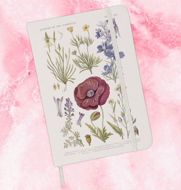 White journal with "Flowers of the Cornfields" illustration, elastic band closure and hanging ribbon bookmark lays on a pink marble surface