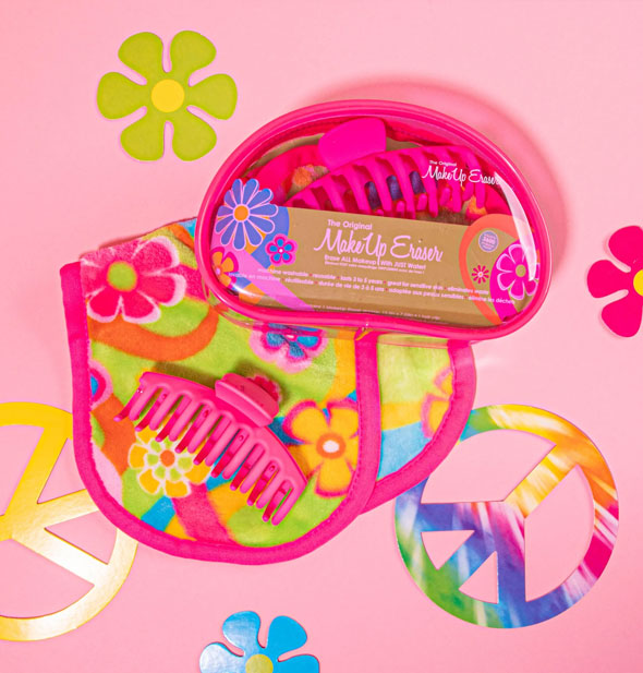 MakeUp Eraser cloth with colorful retro-style floral print lays with a pink claw clip and a clear pouch on a pink surfaced staged with colorful  flowers and peace signs
