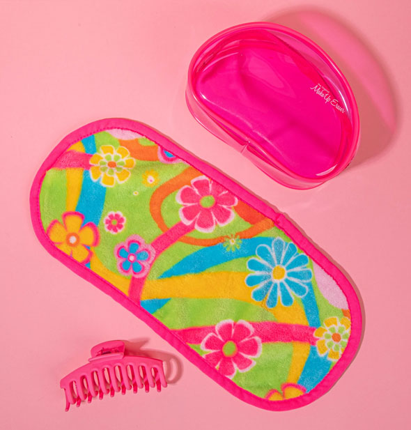 MakeUp Eraser cloth with colorful retro-style floral pattern lays with a pink claw clip and clear pink pouch on a pink surface