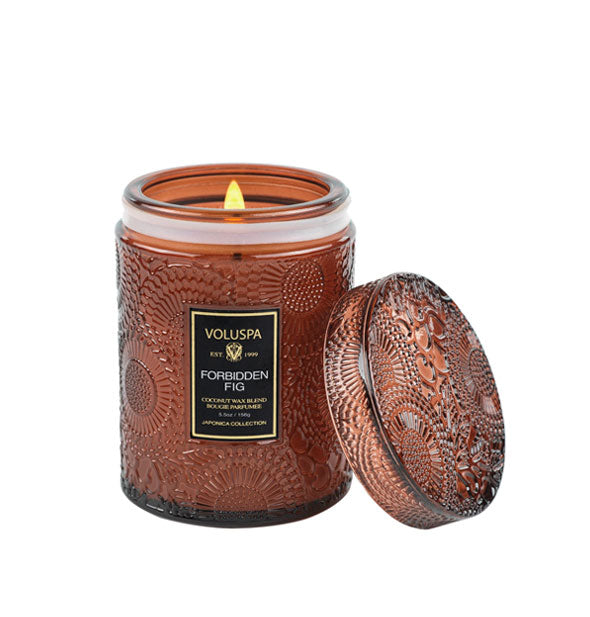 Brown embossed glass candle jar with matching glass lid propped against it features a lit wick and black Voluspa: Forbidden Fig label