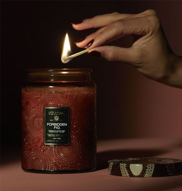 Model's hand holds a lit match above a Voluspa Forbidden Fig candle jar