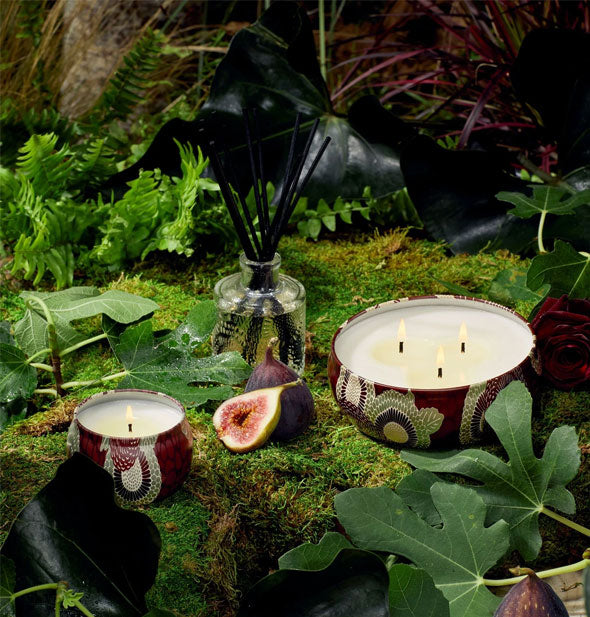 Voluspa candle tins staged with diffuser, figs, and florals on mossy backdrop