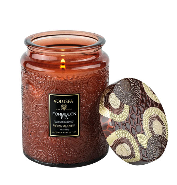 Brown embossed glass candle jar with ornately decorated metal lid propped against it features a lit wick and black Voluspa: Forbidden Fig label