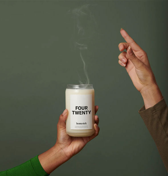 Model's hand holds a smoking Four Twenty candle as another model's hand signals upward with a pointed finger