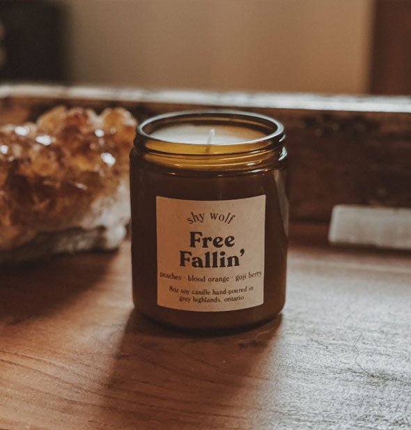 8 ounce amber glass Free Fallin' candle by Shy Wolf sits on a wooden tabletop with raw quartz in the background