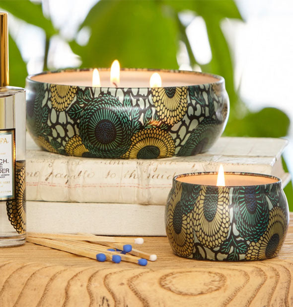 Decorative green and gold tin candles, one with three lit wicks and the other with one lit wick, sit on a wooden surface with matches and books