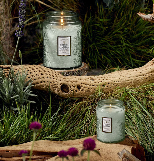 One small and one large green embossed glass Voluspa candle sit among driftwood and purple wildflowers against a grassy backdrop
