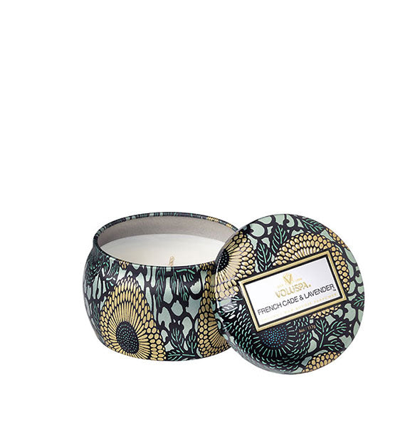 A small unlit candle inside a rounded tin with blue and gold metallic floral design and matching lid set to the side.