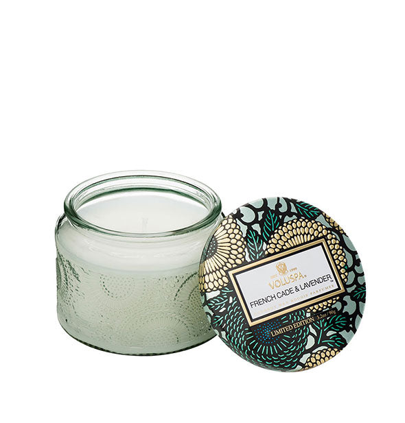 Light green embossed glass French Cade & Lavender Voluspa candle jar with green and gold metallic floral lid set to the side