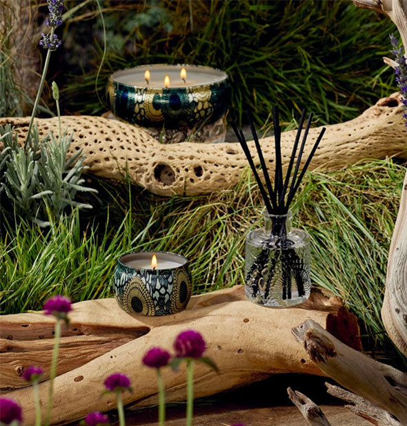 Decorative green and gold tin candles with lit wicks are staged with a reed diffuser, driftwood, and purple wildflowers against a grassy backdrop